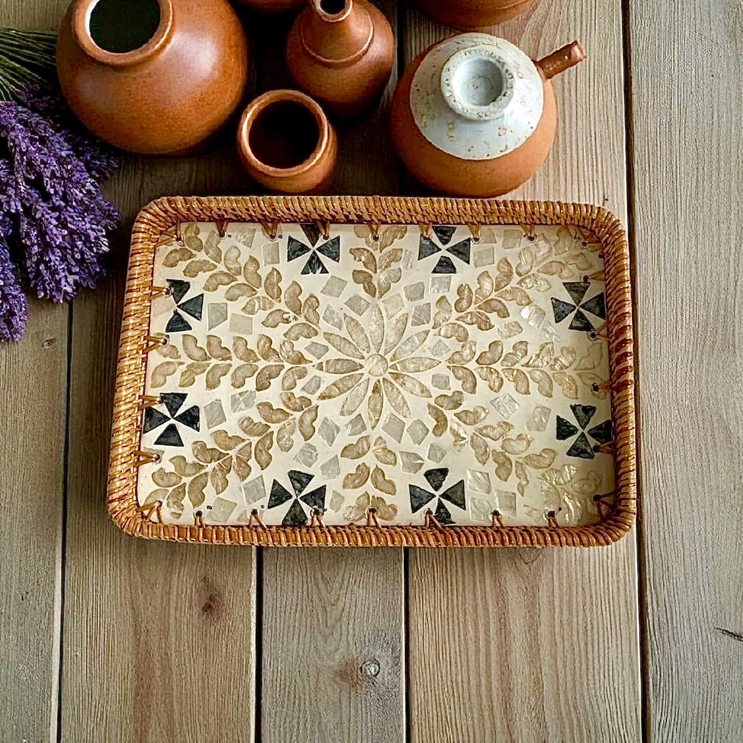Rectangular mother-of-pearl inlay tray with rattan rim on rustic wood beside clay pots; beige floral mosaic.