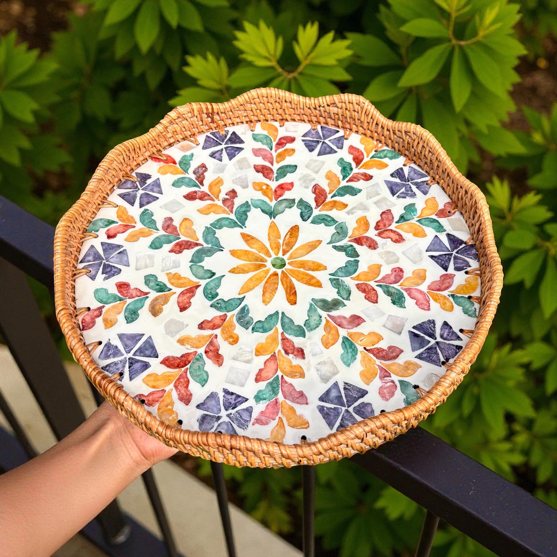 Round woven mat with colorful floral pattern held by a hand against a green leafy background