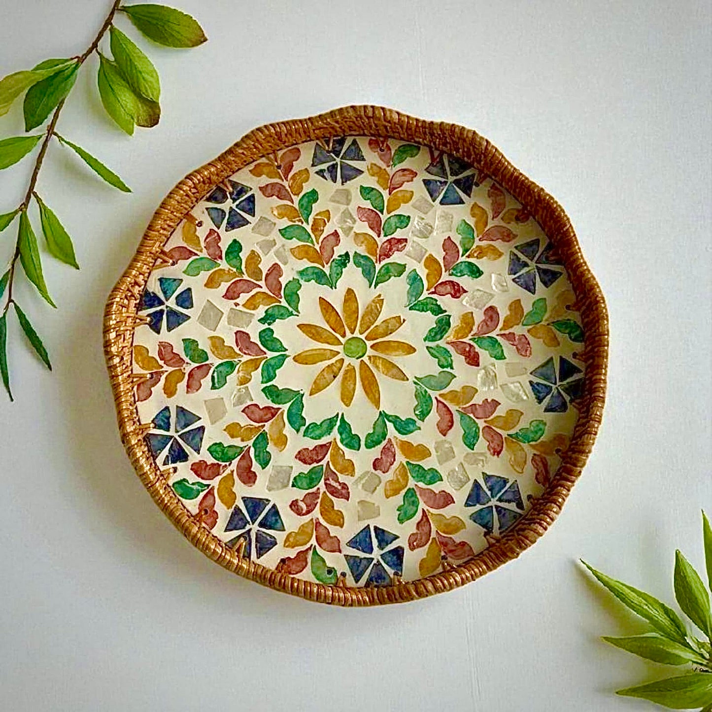 Blossom Scalloped Round Tray with multicolour mother-of-pearl mosaic and rattan rim on a light background with green leaves