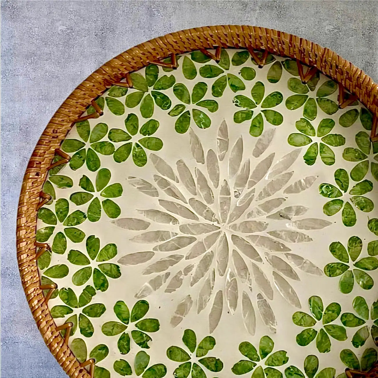 Round mother-of-pearl tray with green floral mosaic and rattan rim on a neutral linen background